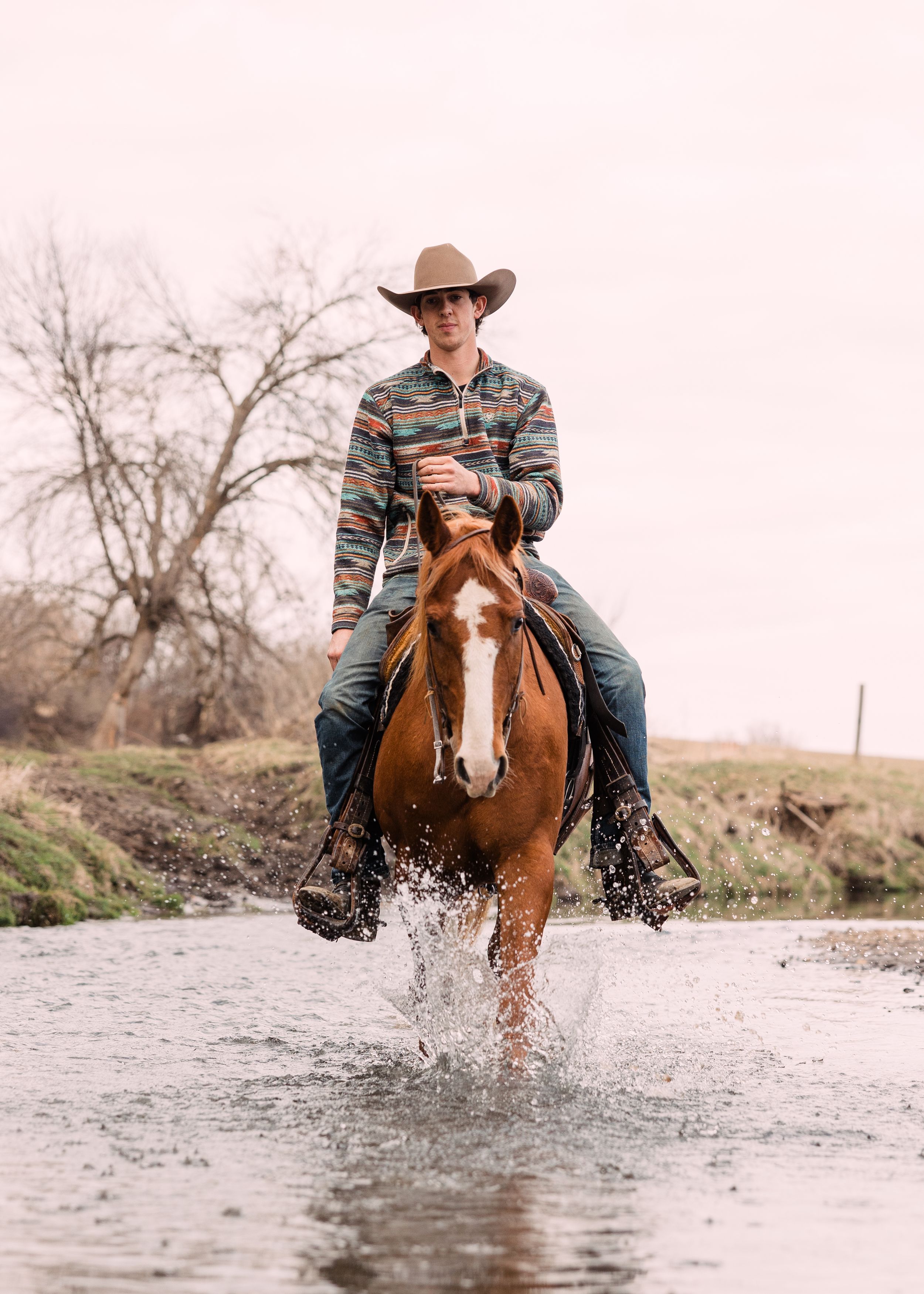A cowboy in a tan hat and striped shirt rides a chestnut horse with a white blaze through shallow, splashing creek water.
