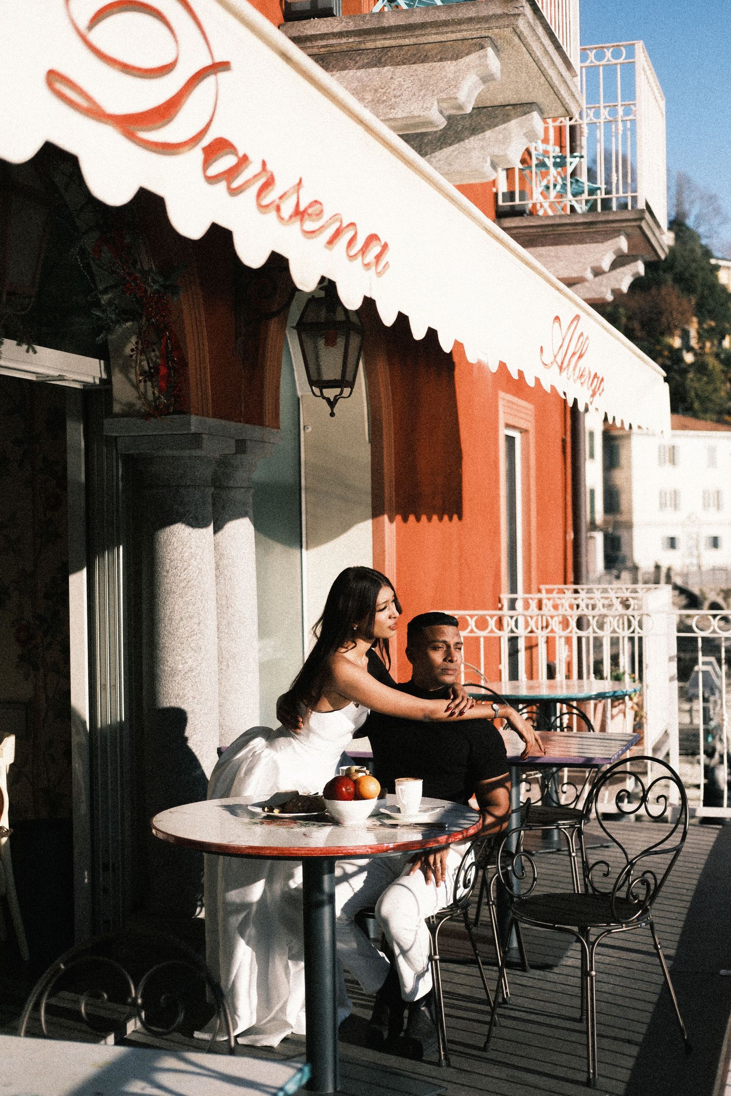 Couple enjoying coffee and snacks at outdoor cafe table overlooking Mediterranean seaside with ornate railing view.