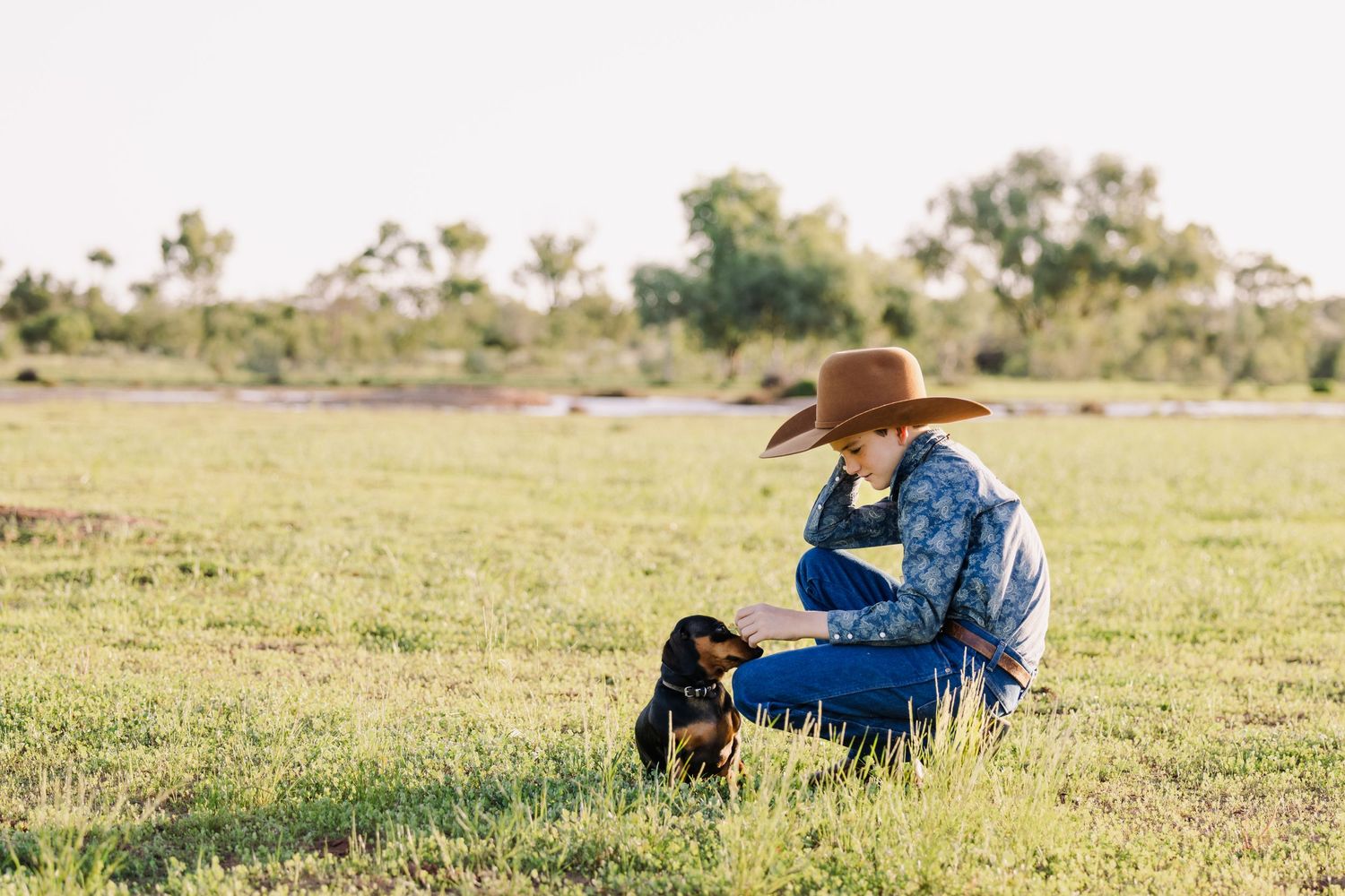 Person in denim and cowboy hat sitting with black dog in grassy field.