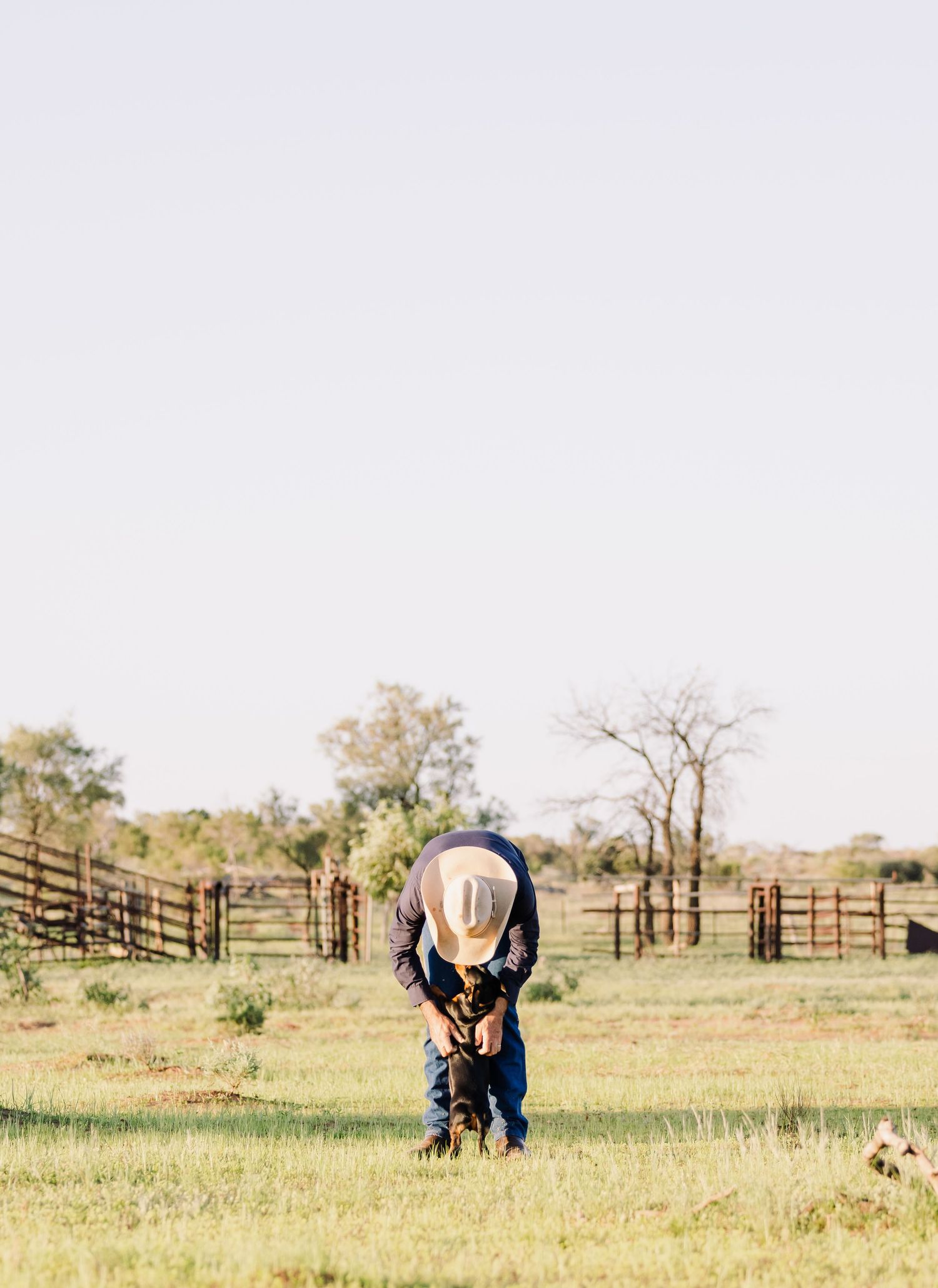Person in denim bending down to pet dog in rural farm setting.