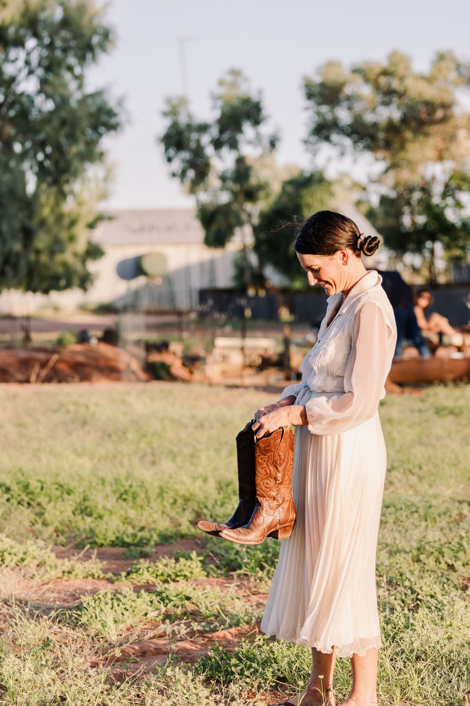 Person in vintage-style dress holding brown leather cowboy boots in rural setting.