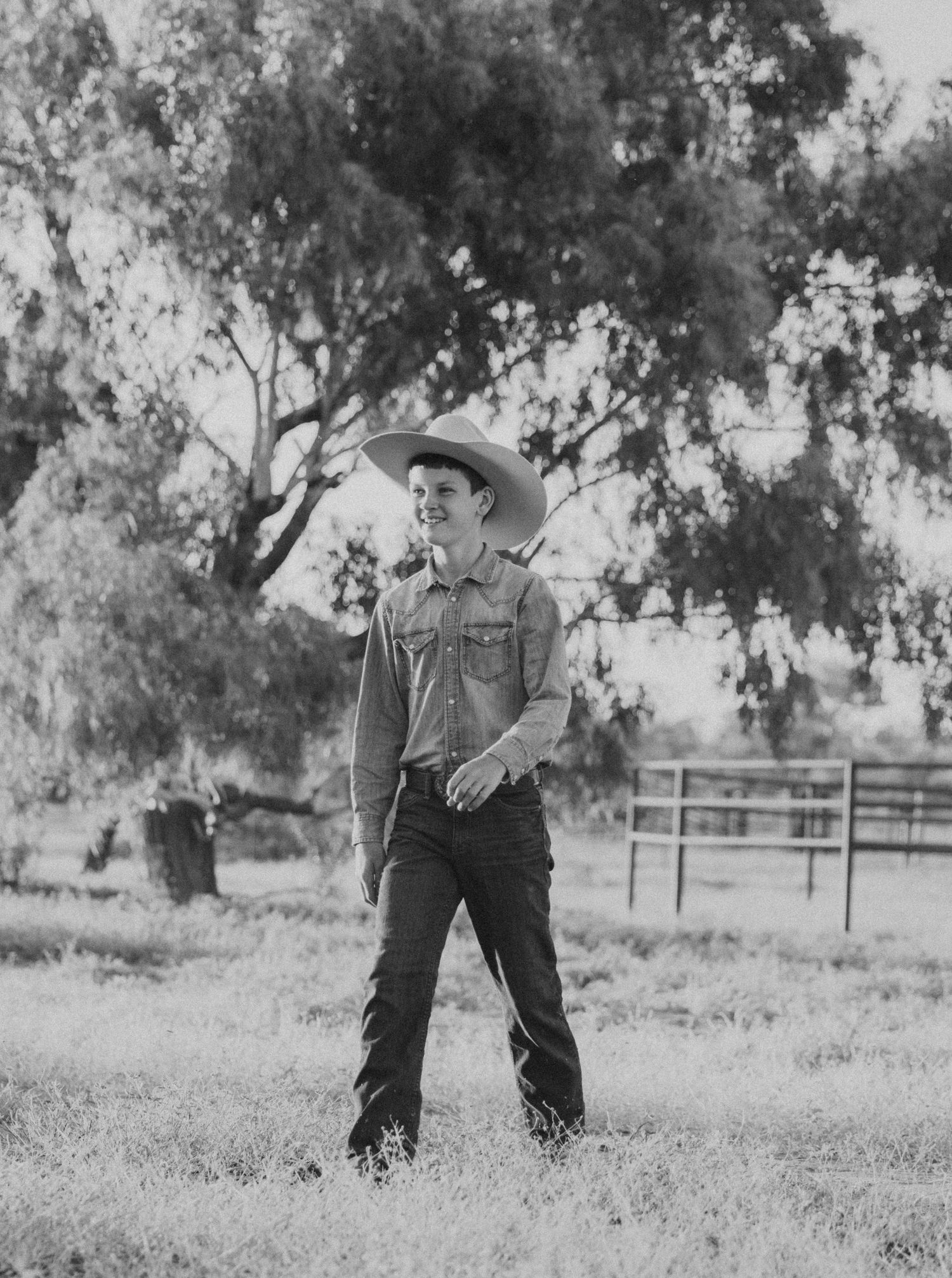 Person in denim and cowboy hat standing in rustic field with trees in black and white photo.