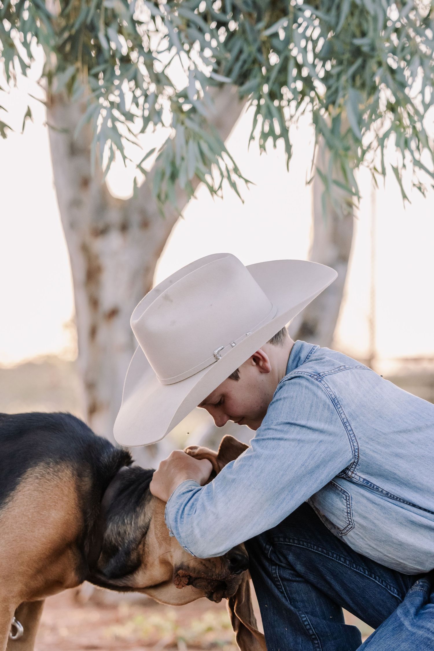 A rancher in a cowboy hat and denim jacket kneels down to pet a black dog under a willow tree.