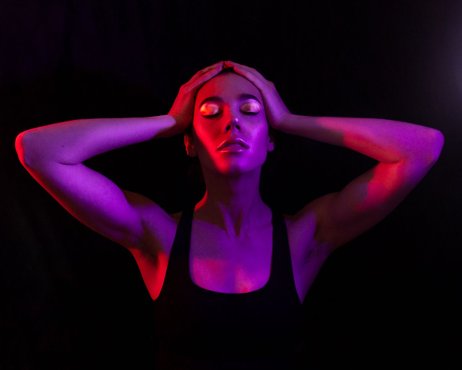 Dramatic portrait with red and purple lighting of a person with hands behind their head.