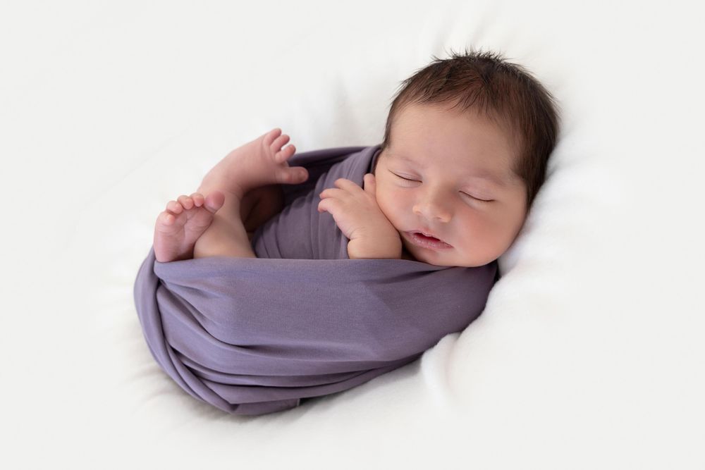 A sleeping newborn wrapped in soft purple fabric against a white background.