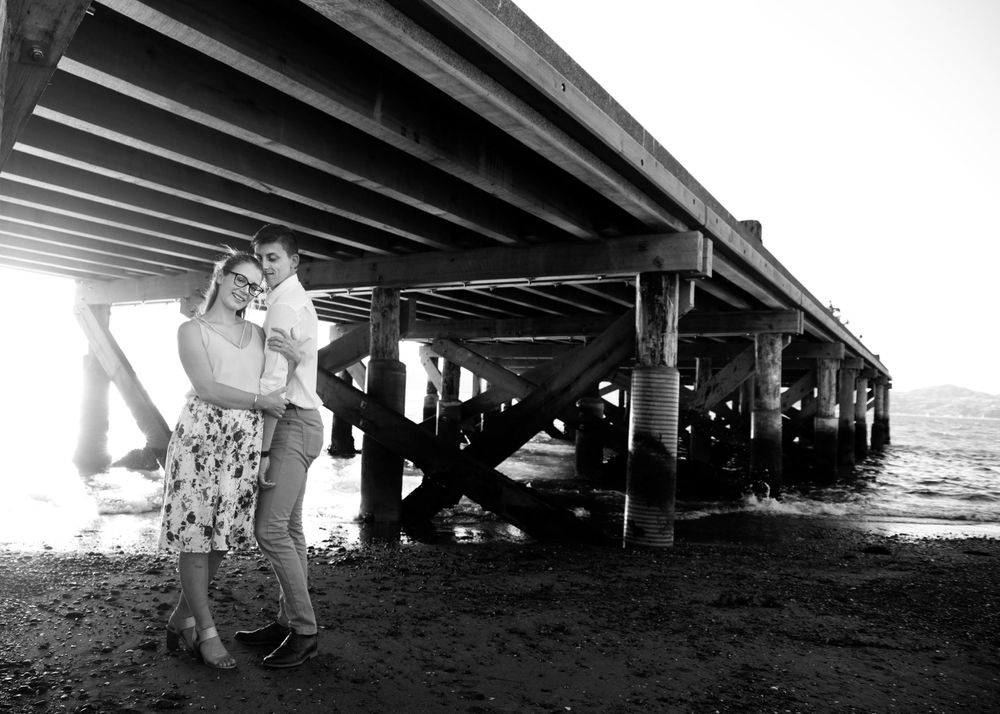 A romantic black and white photo shows a couple embracing underneath a large wooden pier by the ocean.