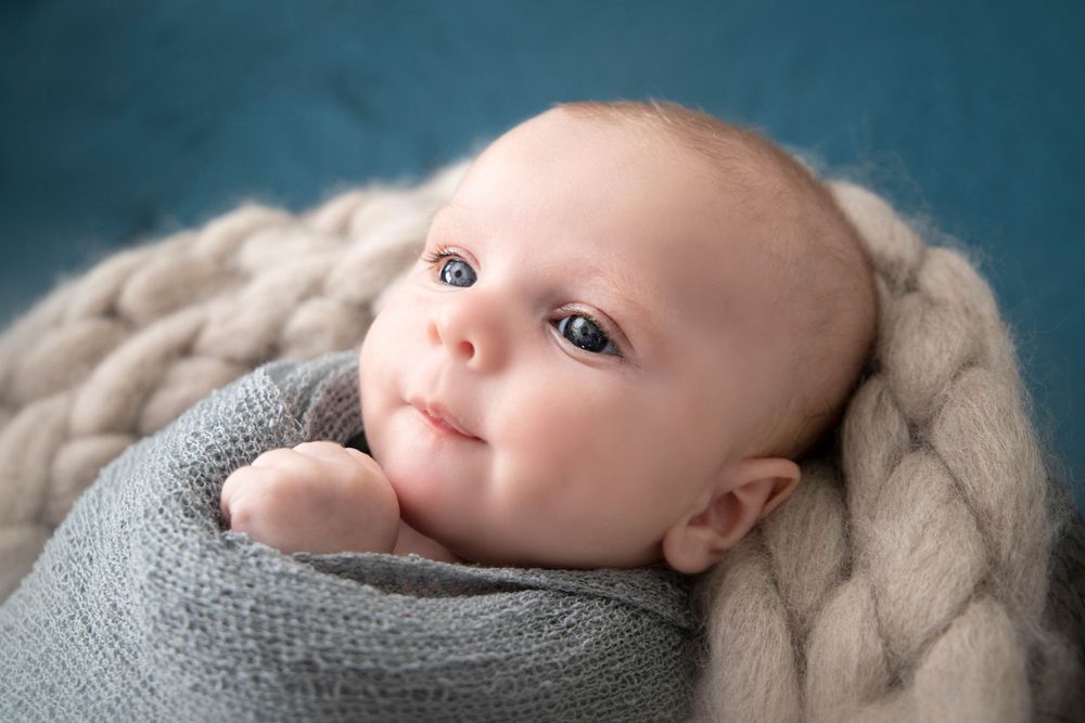 Sweet baby swaddled in gray fabric lying on cream colored knit blanket during professional portrait session.