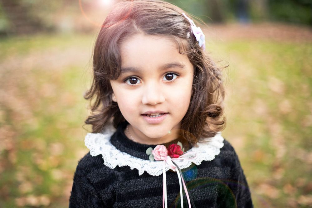 Child wearing black dress with white lace collar and flower hair accessory poses outdoors during autumn.