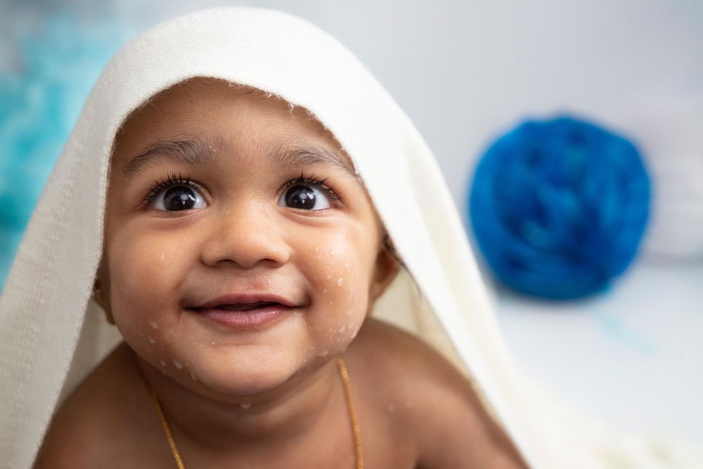 A happy baby wearing a white towel hood smiles while looking upward with a blue sphere in the blurred background.