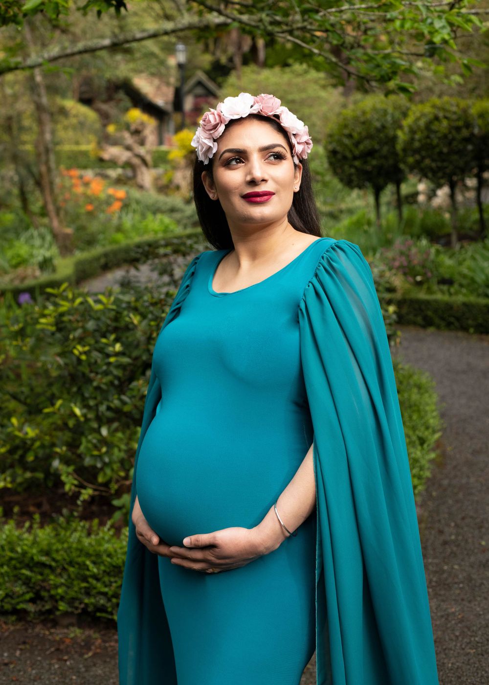An expectant figure in a teal form-fitting dress with cape sleeves and floral crown poses in a lush garden setting.