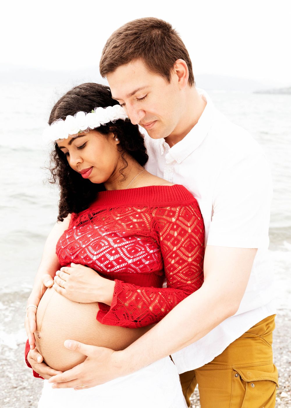 A couple in casual attire embraces on a beach, with a white floral headpiece and red lace top highlighting the maternity pose.