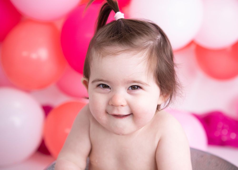 Smiling baby against a pink and red balloon backdrop during a festive photo session.