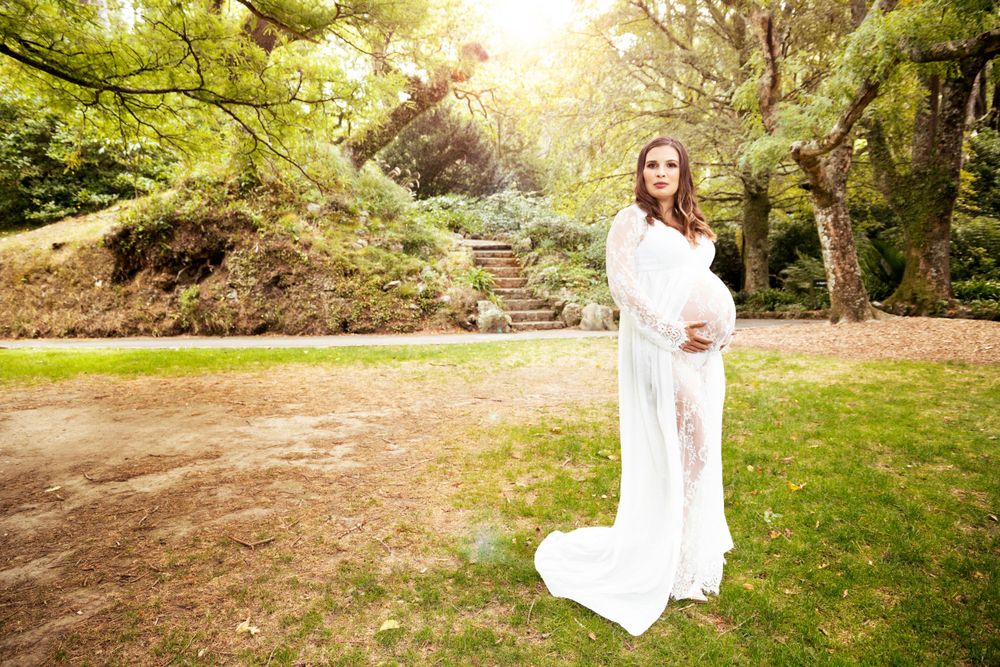 A pregnant person in an elegant white lace gown poses on a lawn near stone steps in a sunlit garden setting.