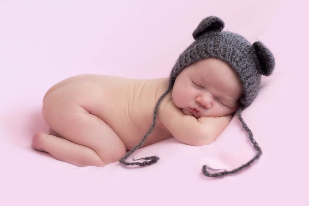Sleeping newborn baby posed on pink background wearing gray knitted bear hat.