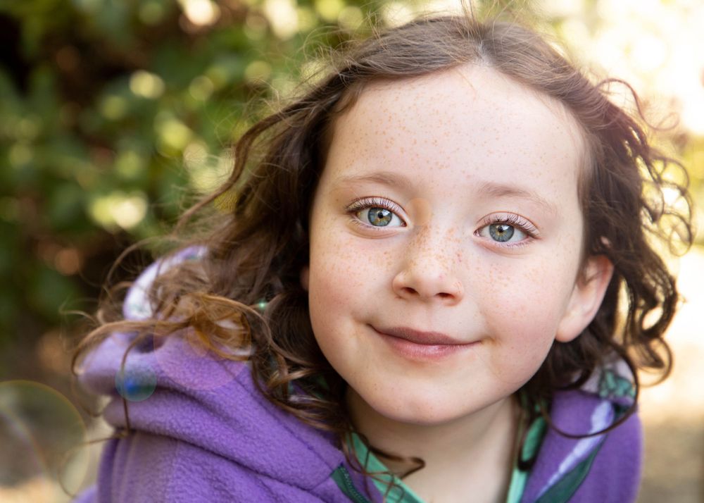 A child wearing a purple jacket smiles at the camera in natural outdoor lighting.