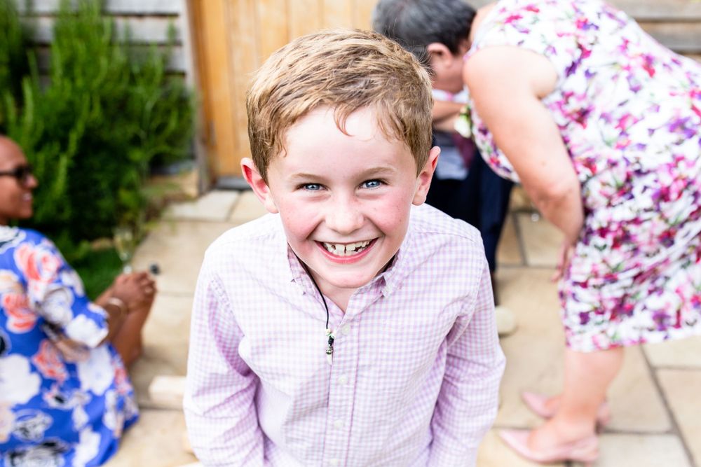 A young child in a pink checkered shirt smiles brightly at the camera while standing outdoors.
