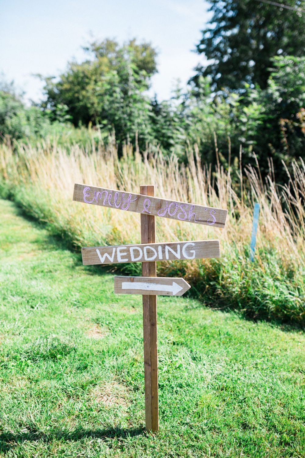 Rustic wooden wedding directional sign with arrows on a post surrounded by tall grass and greenery.