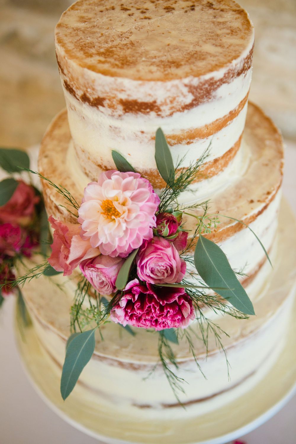 Three-tier semi-naked wedding cake decorated with pink peonies, roses, and eucalyptus leaves on a white cake stand.