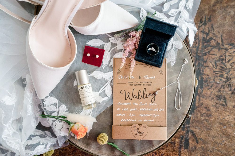 Wedding details on mirrored tray including wooden invitation, earrings, perfume, and white high heels.