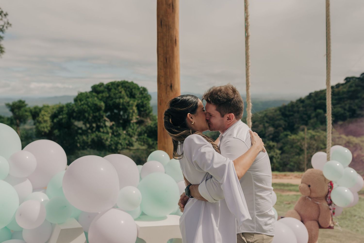 Couple sharing romantic kiss outdoors surrounded by pastel balloons at scenic overlook.