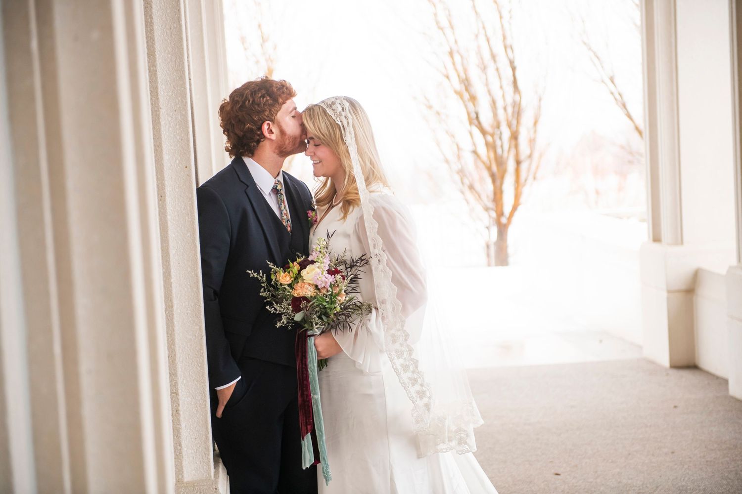 Newlywed couple shares romantic moment on porch with winter scenery in background as bride holds floral bouquet.