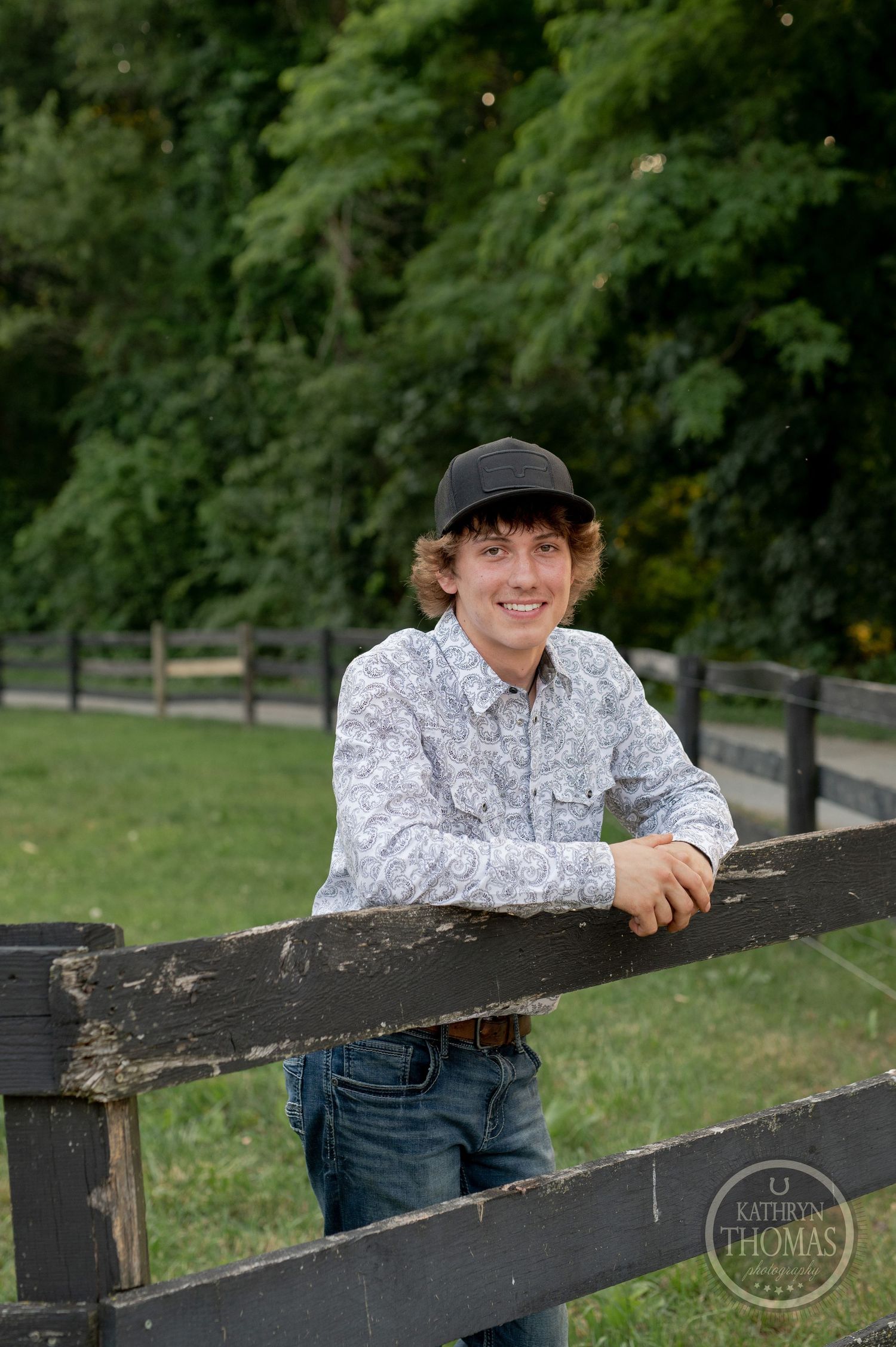 A person wearing a baseball cap leans on a wooden fence with trees in the background.