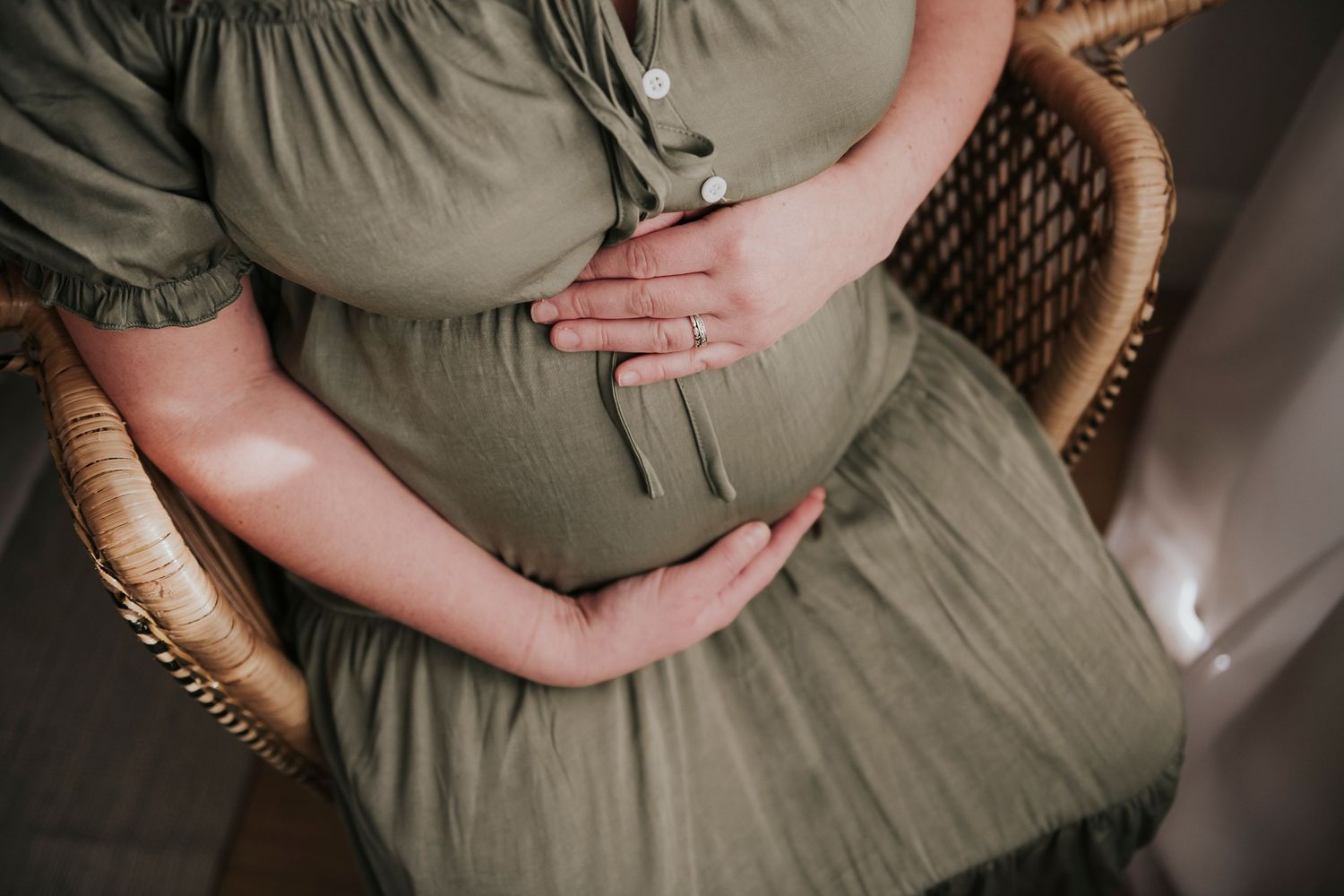 Overhead view of hands cradling a baby bump in olive green maternity dress while sitting in wicker chair.