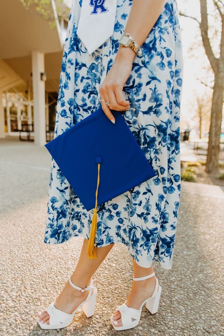 Graduate in blue and white floral dress holds blue graduation cap and wears white strappy heels during outdoor ceremony.