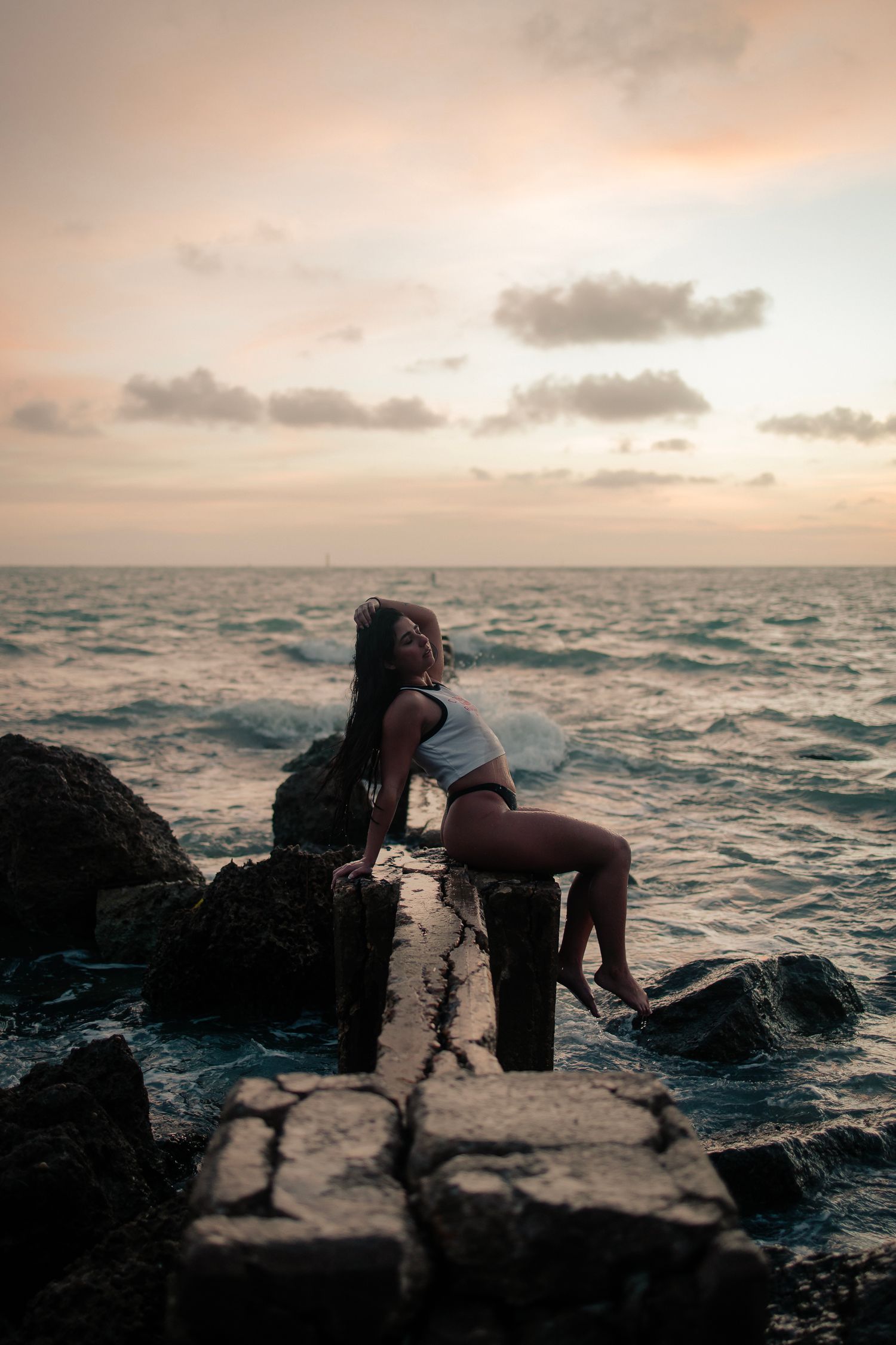 Cinematic beach portrait of woman by ocean rocks at sunset. Mannyportraits South Florida photography with moody, cinematic style.