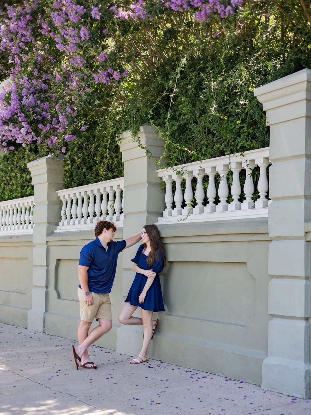 Two people stand by an ornate white balustrade wall with purple flowering vines above it in a garden setting.