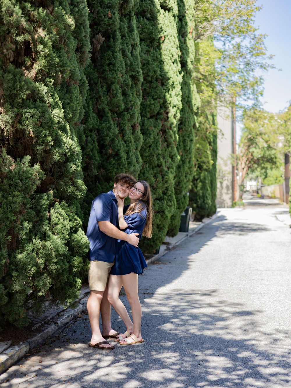 A couple embraces on a scenic tree-lined pathway with tall cypress trees and dappled sunlight creating a romantic scene.
