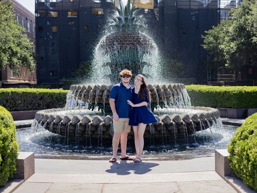 Two people pose in front of the iconic Pineapple Fountain at Charleston's Waterfront Park on a sunny day.
