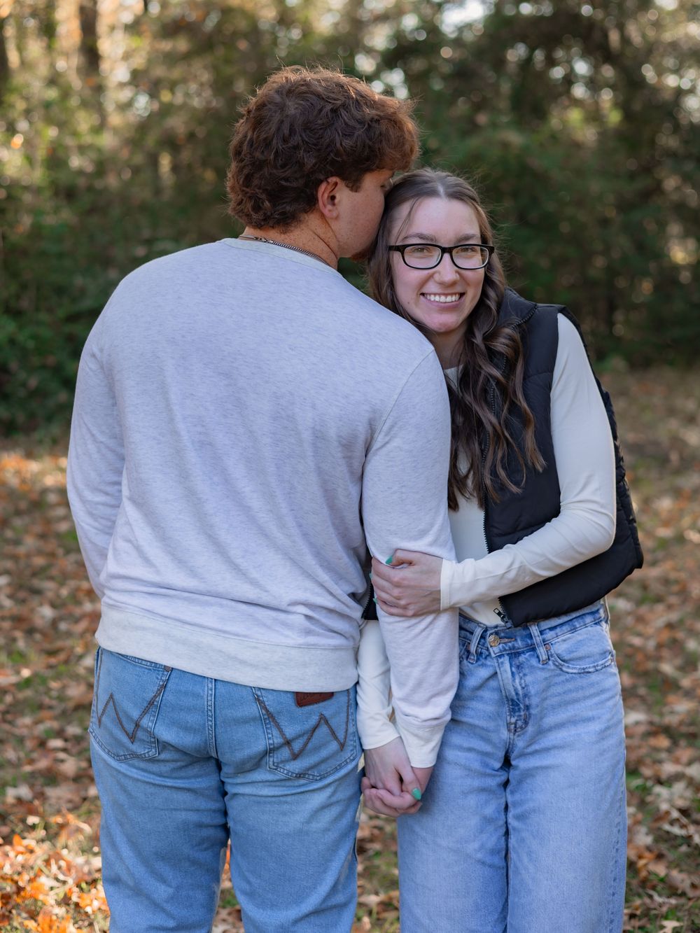 A couple in jeans and sweaters share a tender moment while walking through autumn leaves in an outdoor setting.