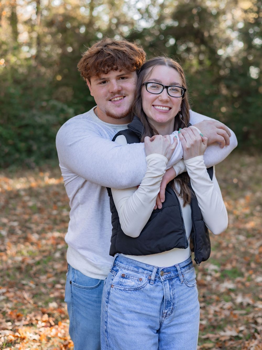 A couple embraces playfully in an autumn forest setting with fallen leaves on the ground.