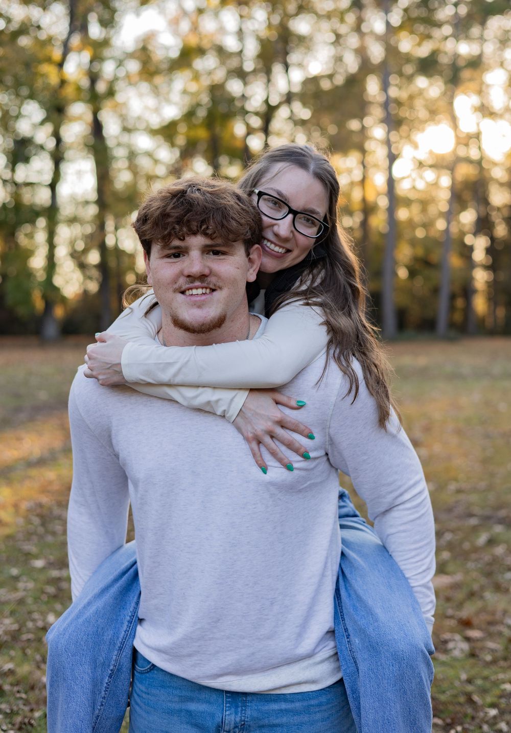 A couple embraces and laughs together outdoors on an autumn day with fallen leaves and trees in the background.