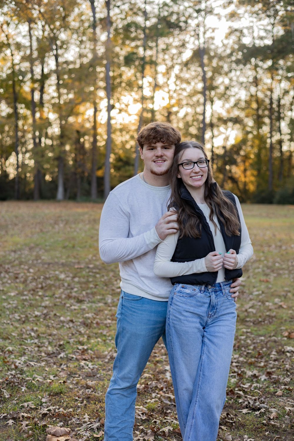 A couple in matching jeans and light tops embrace while standing in a field of autumn leaves at sunset.