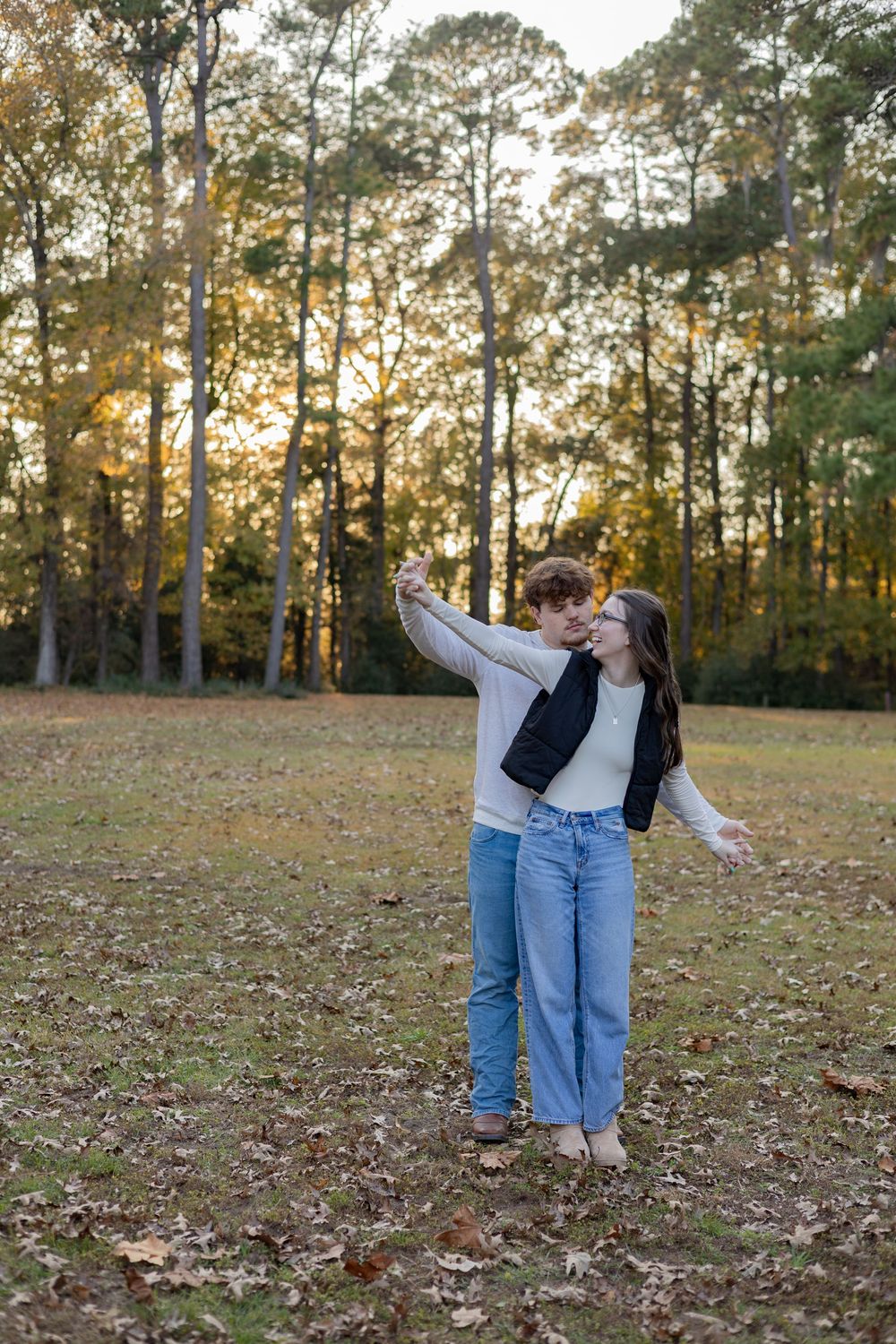 A person in jeans and white shirt dances playfully in a fall forest setting with golden sunlight filtering through trees.