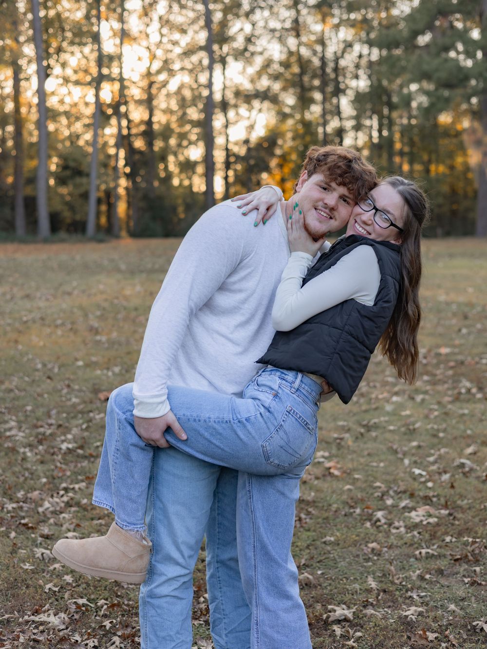 A couple in casual attire playfully dances together on fallen autumn leaves during a golden sunset in a park.
