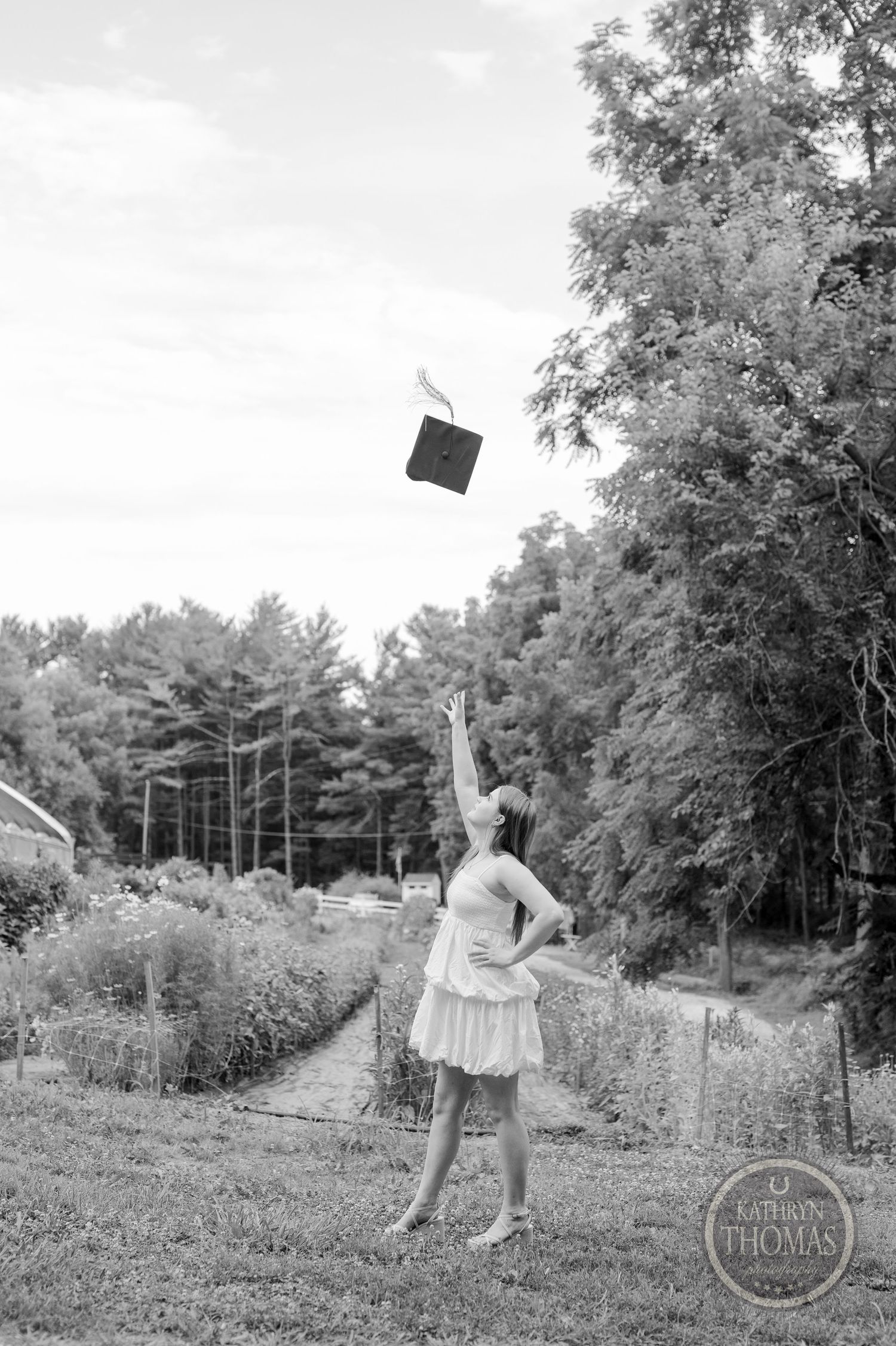 Graduate throws cap in celebration against wooded background in black and white portrait.