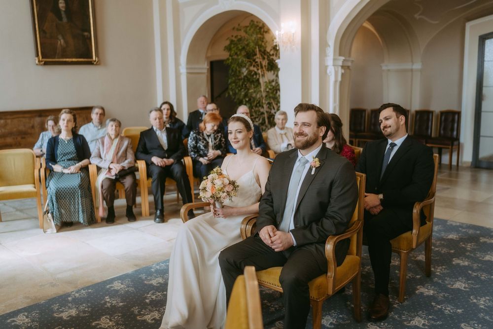 Wedding ceremony guests seated in wooden chairs inside an elegant room with arched doorways and ornate architectural details.