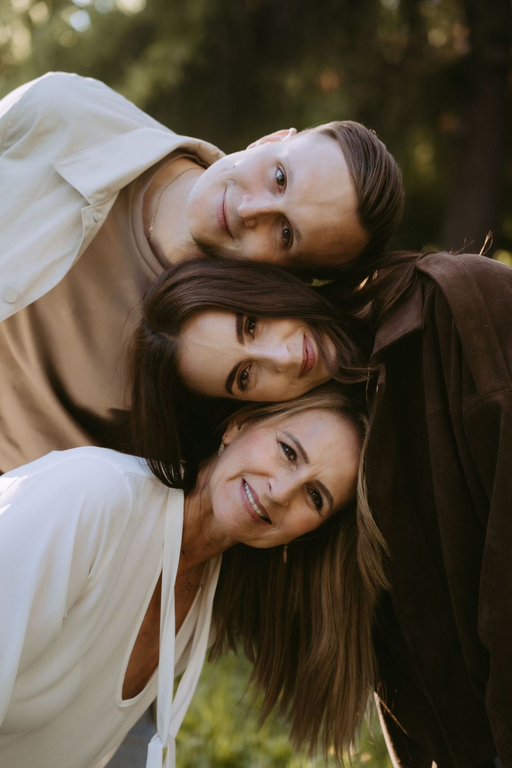 Three young women with brown hair smile warmly while leaning heads together outdoors among green trees.