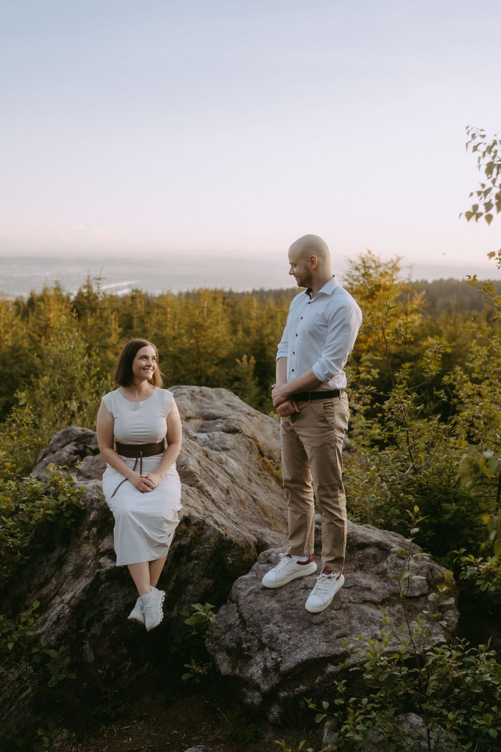 Couple posing on a rocky hilltop surrounded by lush green trees at sunset, woman in white dress, man in white shirt.
