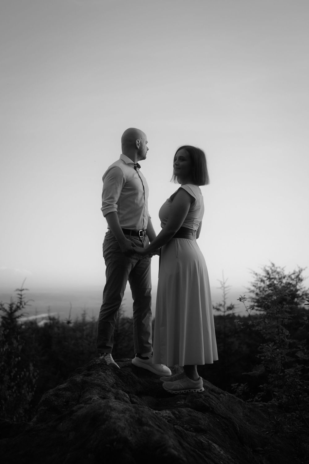 Black and white photo of a couple holding hands on a rocky hilltop at dusk, surrounded by trees and a vast landscape.