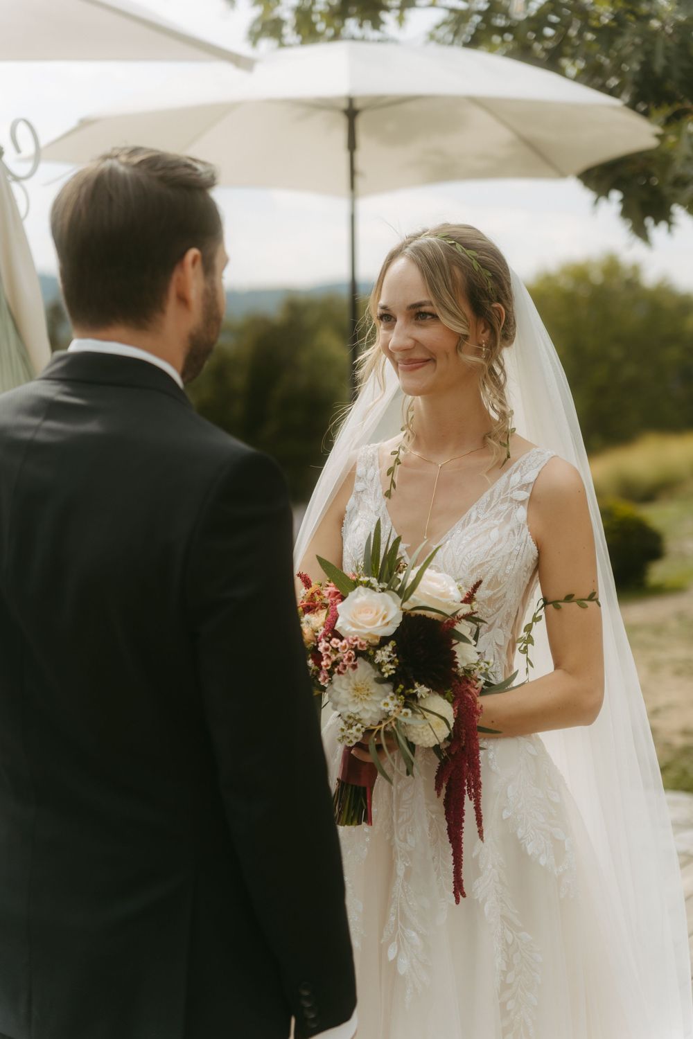 Bride in lace gown holding bouquet smiles at groom during outdoor wedding ceremony under white umbrella.