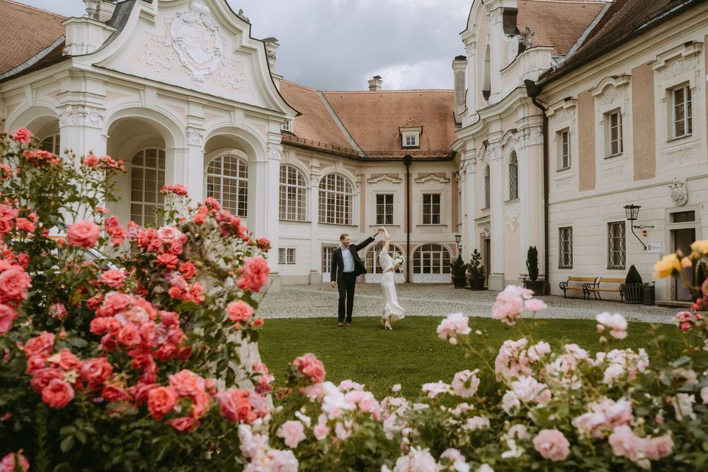 Couple dancing in courtyard of elegant white baroque palace surrounded by blooming pink roses.