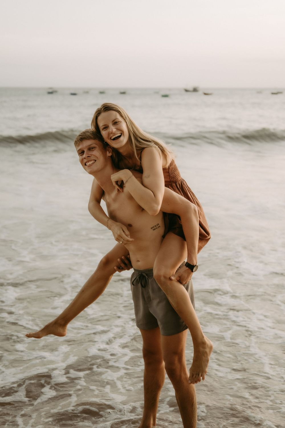 A couple enjoys a playful piggyback ride along the beach at sunset with waves crashing behind them.