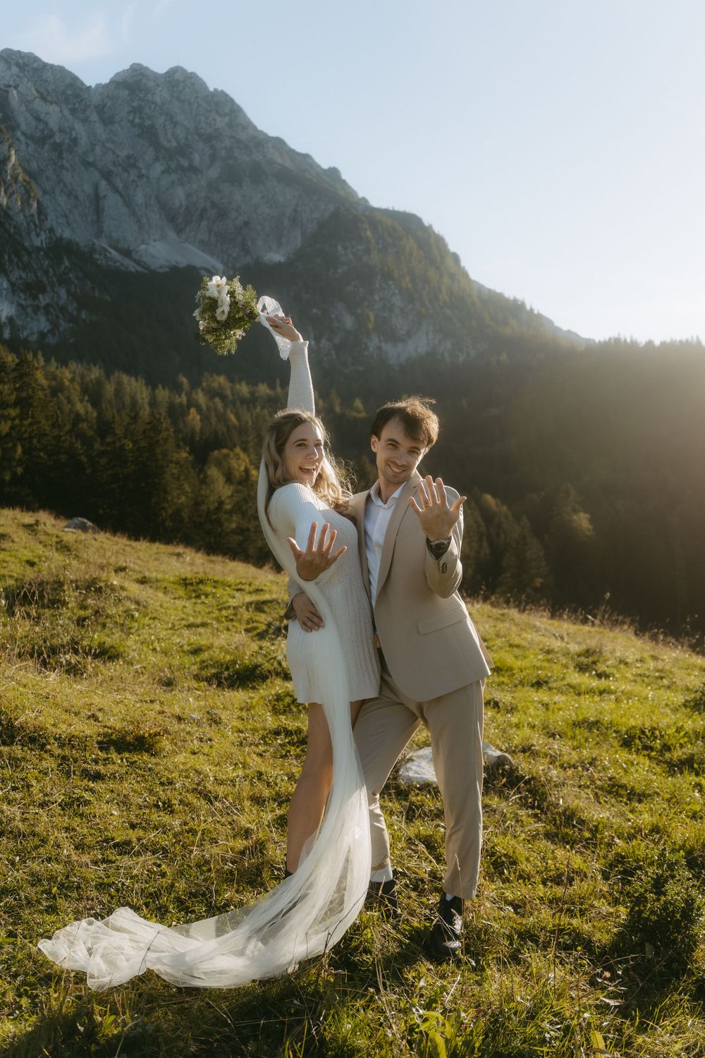 Bride and groom celebrating outdoors on a mountain meadow, showing wedding rings, with rocky peaks and golden sunlight behind them.