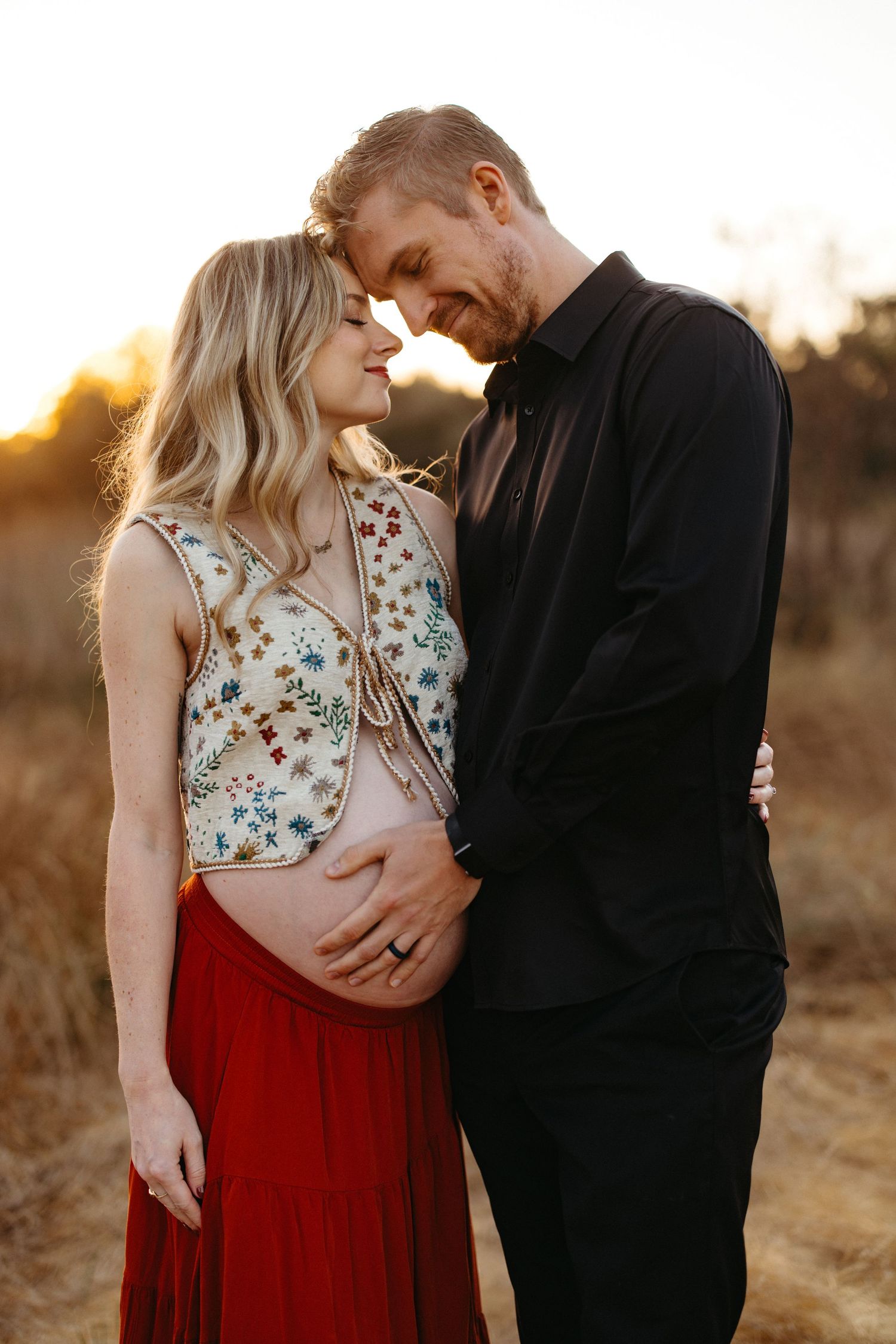 A romantic maternity couple embraces during golden hour sunset in a field wearing black and floral dress.