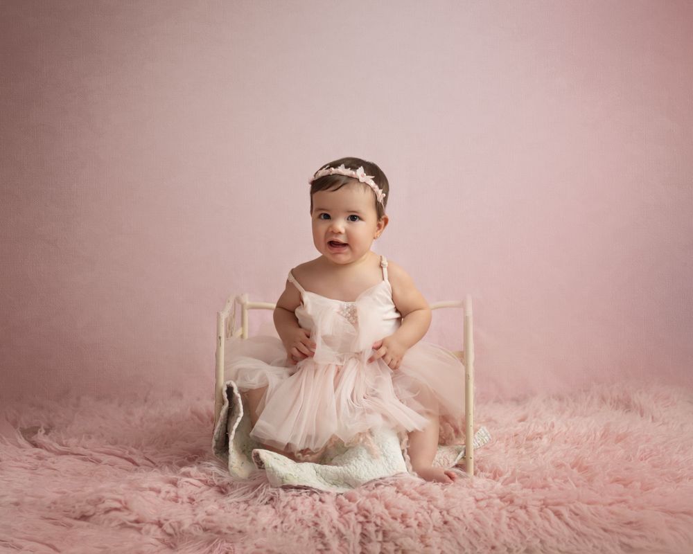 Baby in pink tulle dress poses on plush pink rug with white wooden prop.