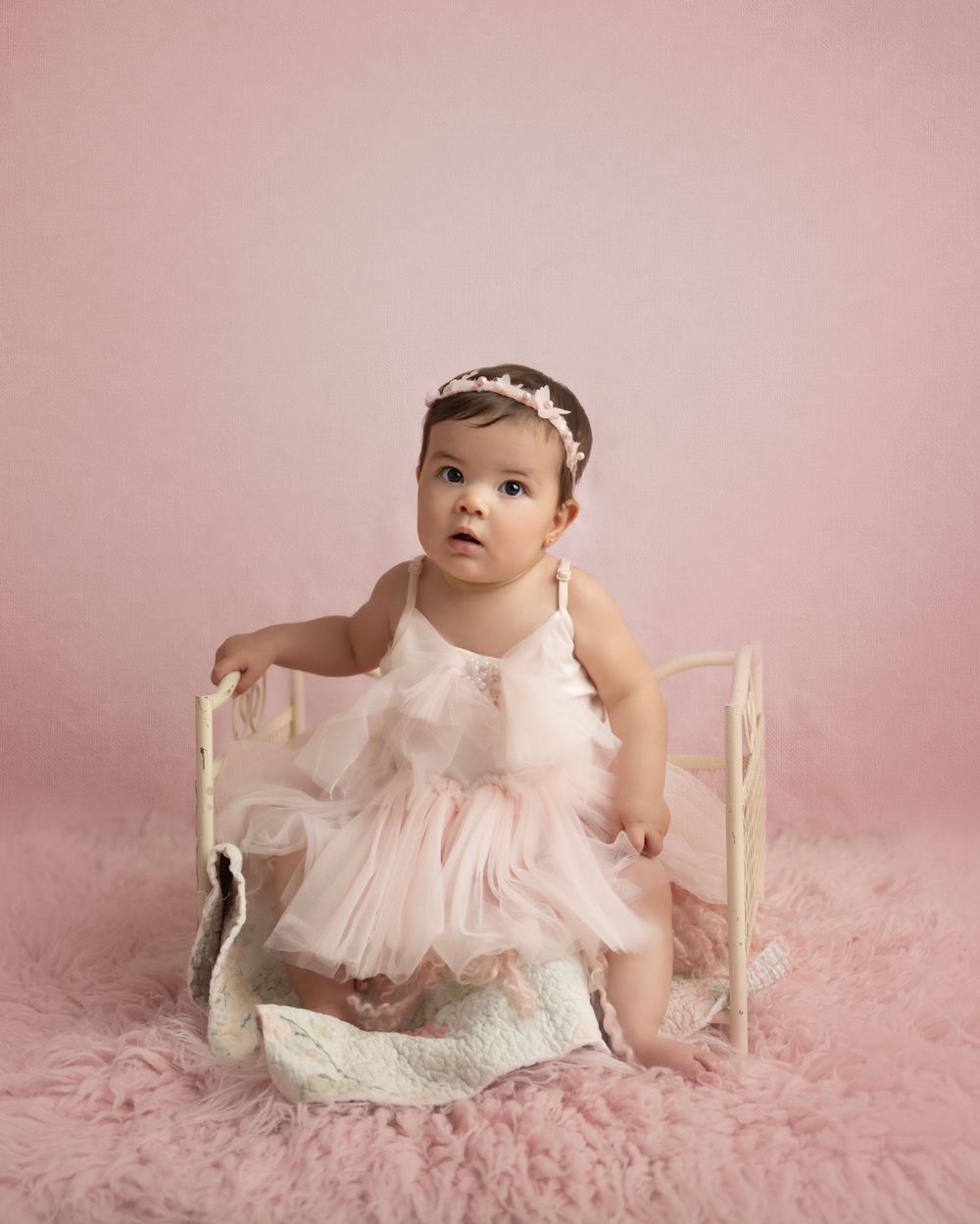 Infant wearing delicate pink tutu dress sits on fluffy pink carpet with white wooden bench.